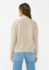 [Color: Heather Cement] A back facing image of a brunette model wearing a beige pullover sweater with a v neckline long sleeves with dropped shoulders. Styles with light wash denim with patch pockets. 
