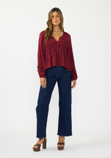 [Color: Merlot] A front facing image of a brunette model wearing a burgundy red long sleeve top with a shirred bust area. Styled with a dark wash denim with patch pockets. 