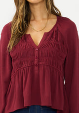 [Color: Merlot] A detail image of a brunette model wearing a burgundy red long sleeve top with a shirred bust area. Styled with a dark wash denim with patch pockets. 