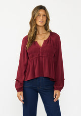 [Color: Merlot] A front facing image of a brunette model wearing a burgundy red long sleeve top with a shirred bust area. Styled with a dark wash denim with patch pockets. 