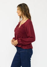 [Color: Merlot] A side facing image of a brunette model wearing a burgundy red long sleeve top with a shirred bust area. Styled with a dark wash denim with patch pockets. 