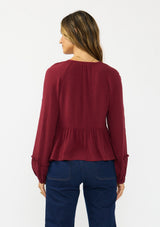 [Color: Merlot] A back facing image of a brunette model wearing a burgundy red long sleeve top with a shirred bust area. Styled with a dark wash denim with patch pockets. 