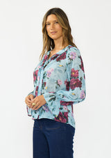[Color: Blue/Burgundy] A side facing image of a brunette model wearing a blue and burgundy floral blouse with a tie neck detail and button front. Styled with dark wash denim. 