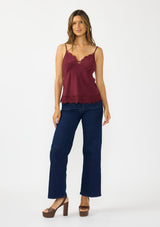 [Color: Merlot] A detail image image of a brunette model wearing a burgundy lace cami with spaghetti straps. Styled with dark wash denim and brown platform heels. 