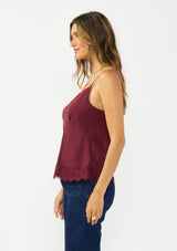 [Color: Merlot] A side facing image of a brunette model wearing a burgundy lace cami with spaghetti straps. Styled with dark wash denim. 