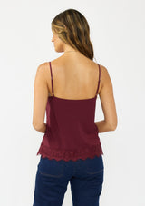 [Color: Merlot] A back facing image of a brunette model wearing a burgundy lace cami with spaghetti straps. Styled with dark wash denim. 