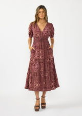 [Color: Wine/Dusty Rose] A front facing image of a brunette model wearing a burgundy bohemian midi dress with  short puff sleeves, a v neckline, elastic waist, and a tiered skirt with ruffle trim. Styled with brown block heels. 