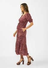 [Color: Wine/Dusty Rose] A side facing image of a brunette model wearing a burgundy bohemian midi dress with  short puff sleeves, a v neckline, elastic waist, and a tiered skirt with ruffle trim. Styled with brown block heels. 