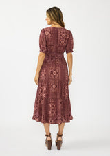 [Color: Wine/Dusty Rose] A back facing image of a brunette model wearing a burgundy bohemian midi dress with  short puff sleeves, a v neckline, elastic waist, and a tiered skirt with ruffle trim. Styled with brown block heels. 