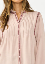 [Color: Mauve] A detail image of a brunette model wearing a pink bohemian blouse with embroidery, a button front, and pintuck detailing. Styled with a dark wash denim with patch pockets. 