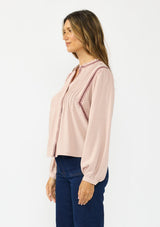 [Color: Mauve] A side facing image of a brunette model wearing a pink bohemian blouse with embroidery, a button front, and pintuck detailing. Styled with a dark wash denim with patch pockets. 
