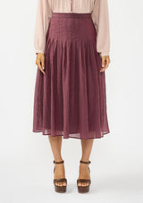 [Color: Plum] A front facing image of a model wearing an embroidered blouse with a crinkled purple midi skirt with pleats. 