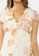 [Color: Ivory/Rust] A detail image of a brunette model wearing a floral maxi dress with short flutter sleeves, a ruffled v neckline, button front, and a front slit. Styled with brown platform heels. 