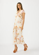 [Color: Ivory/Rust] A side facing image of a brunette model wearing a floral maxi dress with short flutter sleeves, a ruffled v neckline, button front, and a front slit. Styled with brown platform heels. 