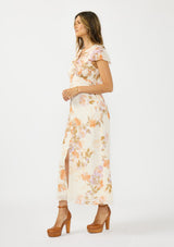 [Color: Ivory/Rust] A side facing image of a brunette model wearing a floral maxi dress with short flutter sleeves, a ruffled v neckline, button front, and a front slit. Styled with brown platform heels. 