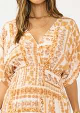 [Color: Taupe/Natural] A detail image of a brunette model wearing a brown floral bohemian maxi dress with short dolman sleeves, a v neckline, smocked waist, and a front slit. 