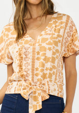 [Color: Taupe/Natural] A detail image of a brunette model wearing a bohemian brown floral top with a v neckline, short sleeves, and a tie front detail.