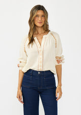[Color: Natural] A front facing image of a brunette model wearing an embroidered blouse with a hidden button front. Styled with a dark wash denim with patch pockets. 