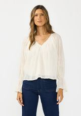 [Color: Vanilla] A front facing image of a brunette model wearing an off white embroidered blouse with sheer sleeves and a ruffle trimmed v neckline. Styled with dark wash patch pocket denim. 