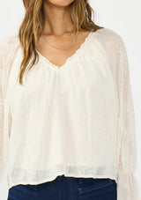 [Color: Vanilla] A detail image of a brunette model wearing an off white embroidered blouse with sheer sleeves and a ruffle trimmed v neckline. Styled with dark wash patch pocket denim. 