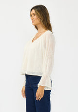 [Color: Vanilla] A side facing image of a brunette model wearing an off white embroidered blouse with sheer sleeves and a ruffle trimmed v neckline. Styled with dark wash patch pocket denim. 