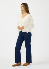 [Color: Vanilla] A side facing image of a brunette model wearing an off white embroidered blouse with sheer sleeves and a ruffle trimmed v neckline. Styled with dark wash patch pocket denim. 