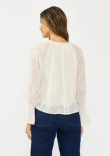 [Color: Vanilla] A back facing image of a brunette model wearing an off white embroidered blouse with sheer sleeves and a ruffle trimmed v neckline. Styled with dark wash patch pocket denim. 
