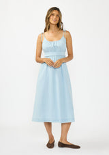[Color: Light Denim Blue] A front facing image of a brunette model wearing a light blue cotton midi dress with tie shoulders and styled with brown suede flats. 