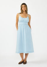[Color: Light Denim Blue] A front facing image of a brunette model wearing a light blue cotton midi dress with tie shoulders and styled with brown suede flats. 