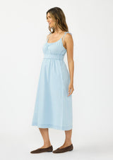 [Color: Light Denim Blue] A side facing image of a brunette model wearing a light blue cotton midi dress with tie shoulders and styled with brown suede flats. 