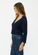 [Color: Navy] A side facing image of a brunette model wearing a navy cropped knit cardigan with sequins and a button front. 