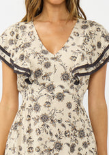 [Color: Natural/Olive] A detail image of a brunette model wearing a beige and black floral print midi dress with short flutter sleeves, a v neckline, and a ruffle trimmed hem. 