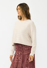 [Color: Birch] A side facing image of a brunette model wearing an off white chenille sweater with a round neck and long sleeves with dropped shoulders. Styled with the burgundy paisley  bohemian maxi skirt. 