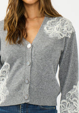 [Color: Heather Grey] A detail image of a brunette model wearing a grey cardigan with lace details, a v neckline, and a button front. 