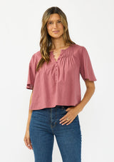 [Color: Rose] A front facing image of a brunette model wearing a pink short sleeve top with a button front and pintucks. Styles with a dark wash denim. 