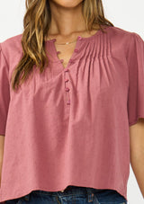 [Color: Rose] A detail image of a brunette model wearing a pink short sleeve top with a button front and pintucks. Styles with a dark wash denim. 