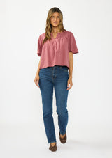 [Color: Rose] A detail image of a brunette model wearing a pink short sleeve top with a button front and pintucks. Styles with a dark wash denim and brown suede ballet flats. 