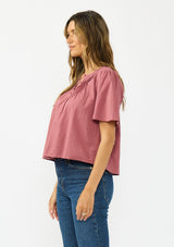 [Color: Rose] A side facing image of a brunette model wearing a pink short sleeve top with a button front and pintucks. Styles with a dark wash denim.