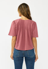 [Color: Rose] A back facing image of a brunette model wearing a pink short sleeve top with a button front and pintucks. Styles with a dark wash denim.