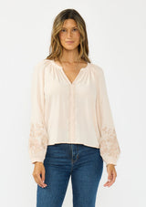 [Color: Petal] A front facing image  of a brunette model wearing a light pink embroidered blouse with a split v neckline. 