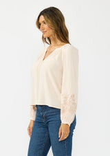 [Color: Petal] A side facing image  of a brunette model wearing a light pink embroidered blouse with a split v neckline. 