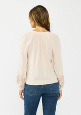 [Color: Petal] A back facing image  of a brunette model wearing a light pink embroidered blouse with a split v neckline. 
