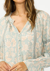 [Color: Dusty Sage/Natural] A detail image of a brunette model wearing a mint floral blouse with a button front, long sleeves, flared elastic cuffs, and lace trims details. 