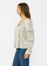 [Color: Dusty Sage/Natural] A side facing image of a brunette model wearing a mint floral blouse with a button front, long sleeves, flared elastic cuffs, and lace trims details. 