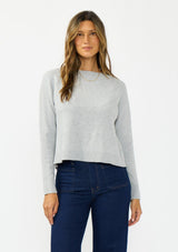 [Color: Heather Silver] A front facing image of a brunette model wearing a grey pullover sweater with a crew neckline and a open back detail. Styled with a dark wash denim with patch pockets. 