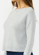 [Color: Heather Silver] A side facing image of a brunette model wearing a grey pullover sweater with a crew neckline and a open back detail. Styled with a dark wash denim with patch pockets. 