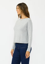 [Color: Heather Silver] A side facing image of a brunette model wearing a grey pullover sweater with a crew neckline and a open back detail. Styled with a dark wash denim with patch pockets. 