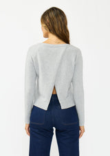 [Color: Heather Silver] A back facing image of a brunette model wearing a grey pullover sweater with a crew neckline and a open back detail. Styled with a dark wash denim with patch pockets. 