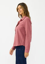 [Color: Rose] A side facing image of a brunette model wearing a pink shirt with a classic collar and button front. Styled with a dark wash denim with patch pockets. 