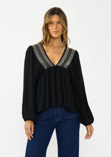 [Color: Black] A front facing image of a brunette model wearing a black long-sleeve blouse with a V-neckline and smocked trim, paired with a dark wash denim.
